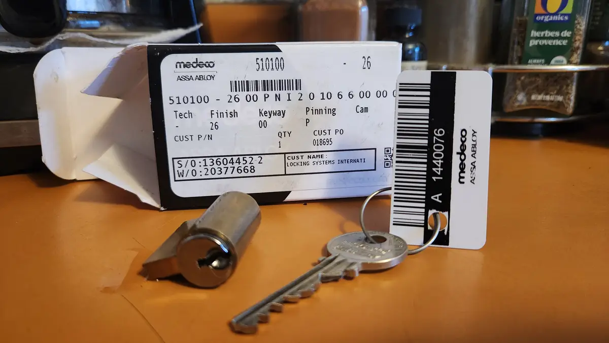 A close-up photo shows a Medeco lock cylinder, its matching key on a keyring, and the original packaging on a wooden…