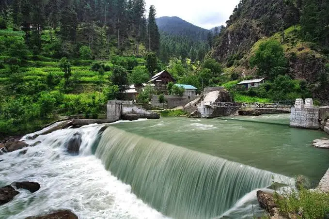 A scenic photo of a river flowing over a small dam in a lush green valley with houses nestled among trees. The image has…