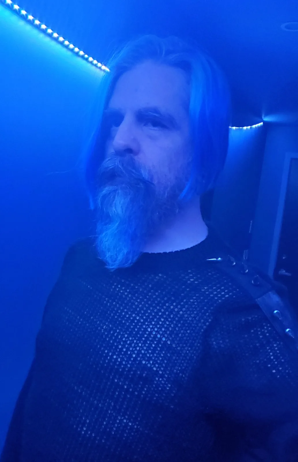 A man with long hair and a beard is shown in a room bathed in blue light from LED strips along the ceiling. He wears a dark…
