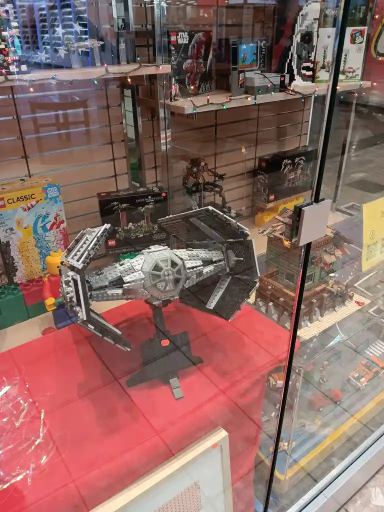 A large, assembled LEGO Star Wars TIE fighter model is displayed on a red cloth inside a glass storefront window, surrounded…