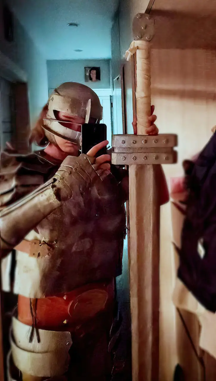 A person in homemade medieval armor, including a helmet and gauntlets, takes a mirror selfie while holding a large sword…