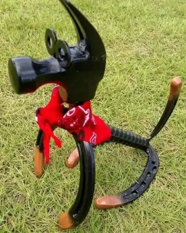 A whimsical sculpture of a dog made from a black hammer head for the head, horseshoes for legs and tail, and a red bandana…