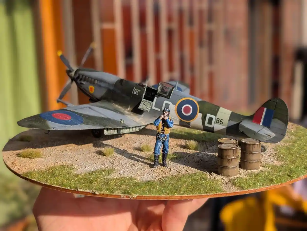 A hand holds a detailed diorama featuring a miniature World War II-era British Spitfire fighter plane with RAF roundels and…