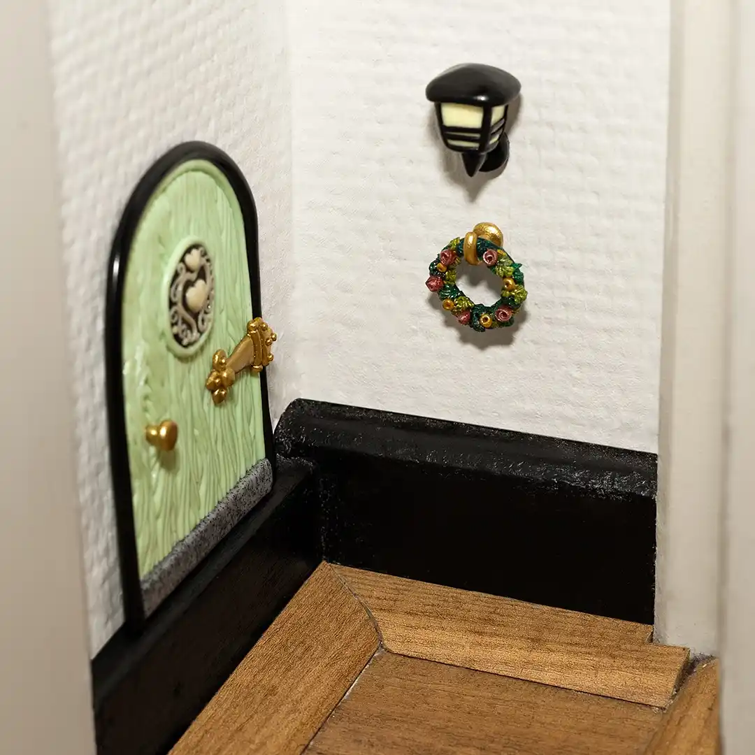 A close-up of a miniature dollhouse scene showing a small green arched door with gold hardware, a tiny black wall-mounted…