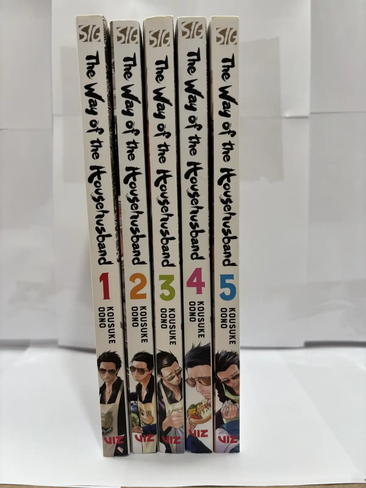 Five manga volumes of 'The Way of the Househusband' by Kou suke Oono stand upright in a row, showing their spines with…