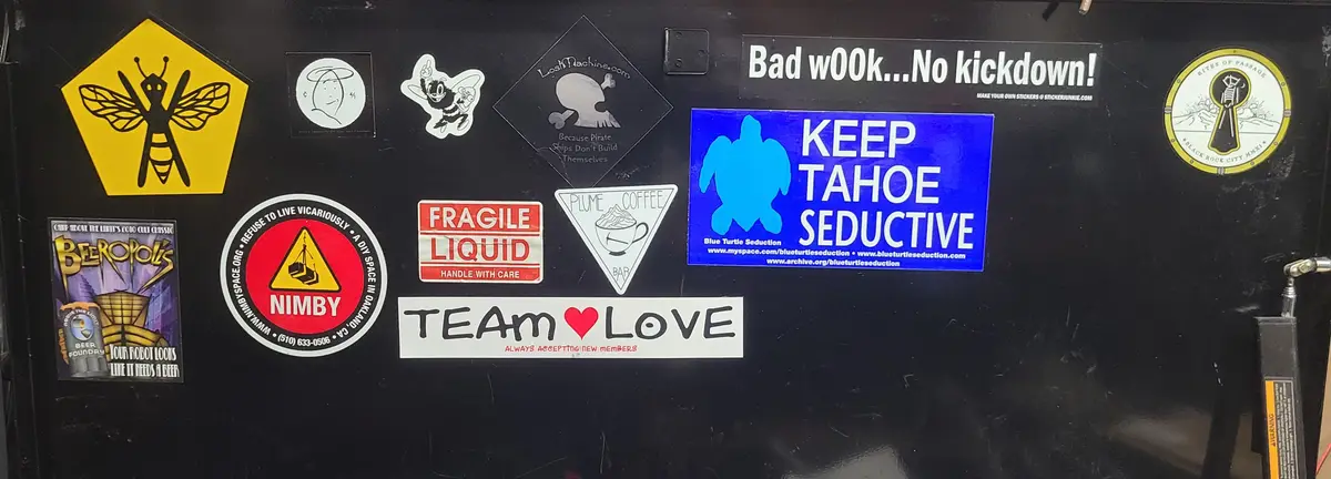 A black surface covered in various stickers, including a yellow bee warning sign, a 'NIMBY' circular sticker, a 'FRAGILE…