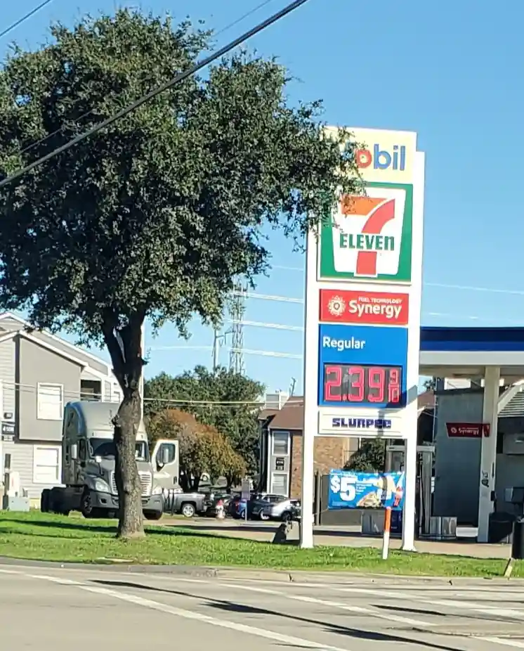 A photo of a Mobil 7-Eleven gas station sign showing the price for regular gas at $2.39 per gallon, with a large tree…