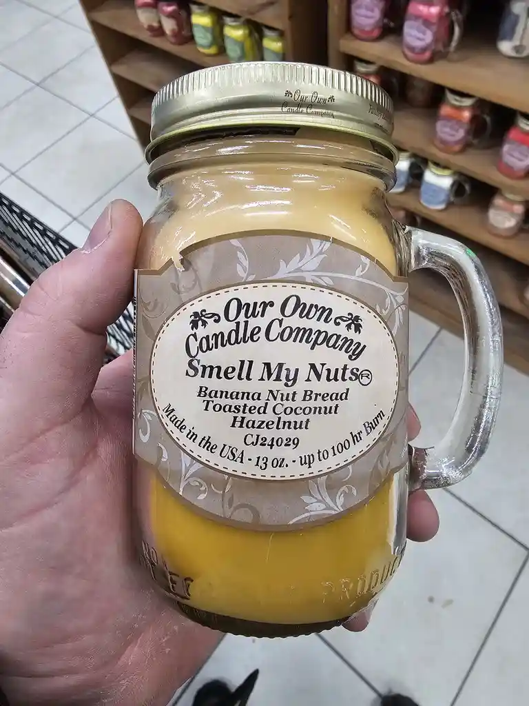 A hand holds a mason jar candle with a label reading 'Our Own Candle Company' and the scent name 'Smell My Nuts®', listing…