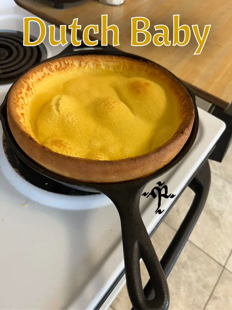 A golden-brown, puffy Dutch baby pancake sits in a black cast iron skillet resting on a white electric stovetop. The text…