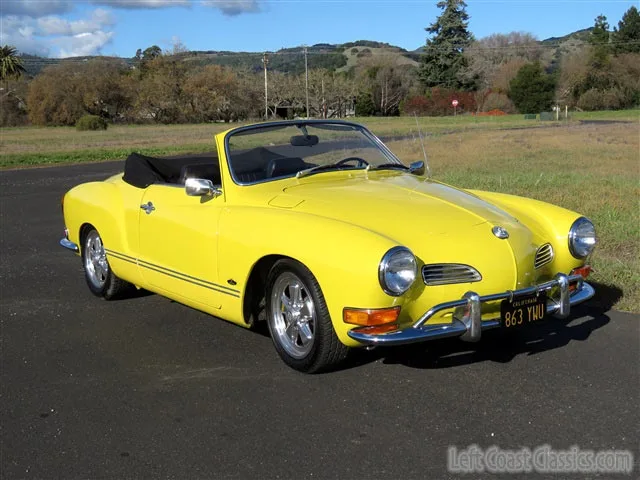 A bright yellow vintage Volkswagen Karmann Ghia convertible is parked on asphalt with a grassy field and hills in the…
