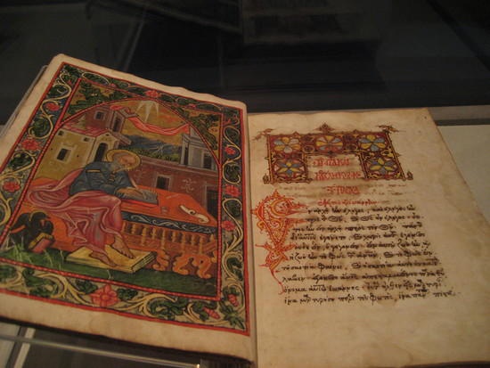 An open, illuminated medieval manuscript with a colorful, detailed illustration on the left page showing a figure writing…