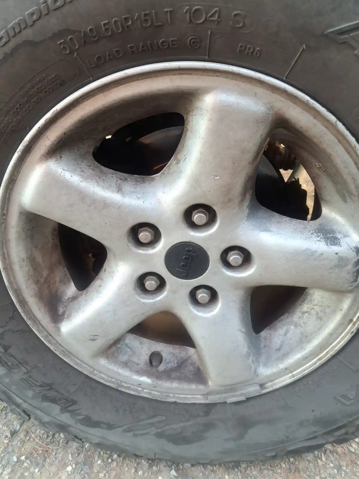 A close-up photo of a dirty, silver Jeep wheel with a visible tire tread and lug nuts, featuring the Jeep logo in the…