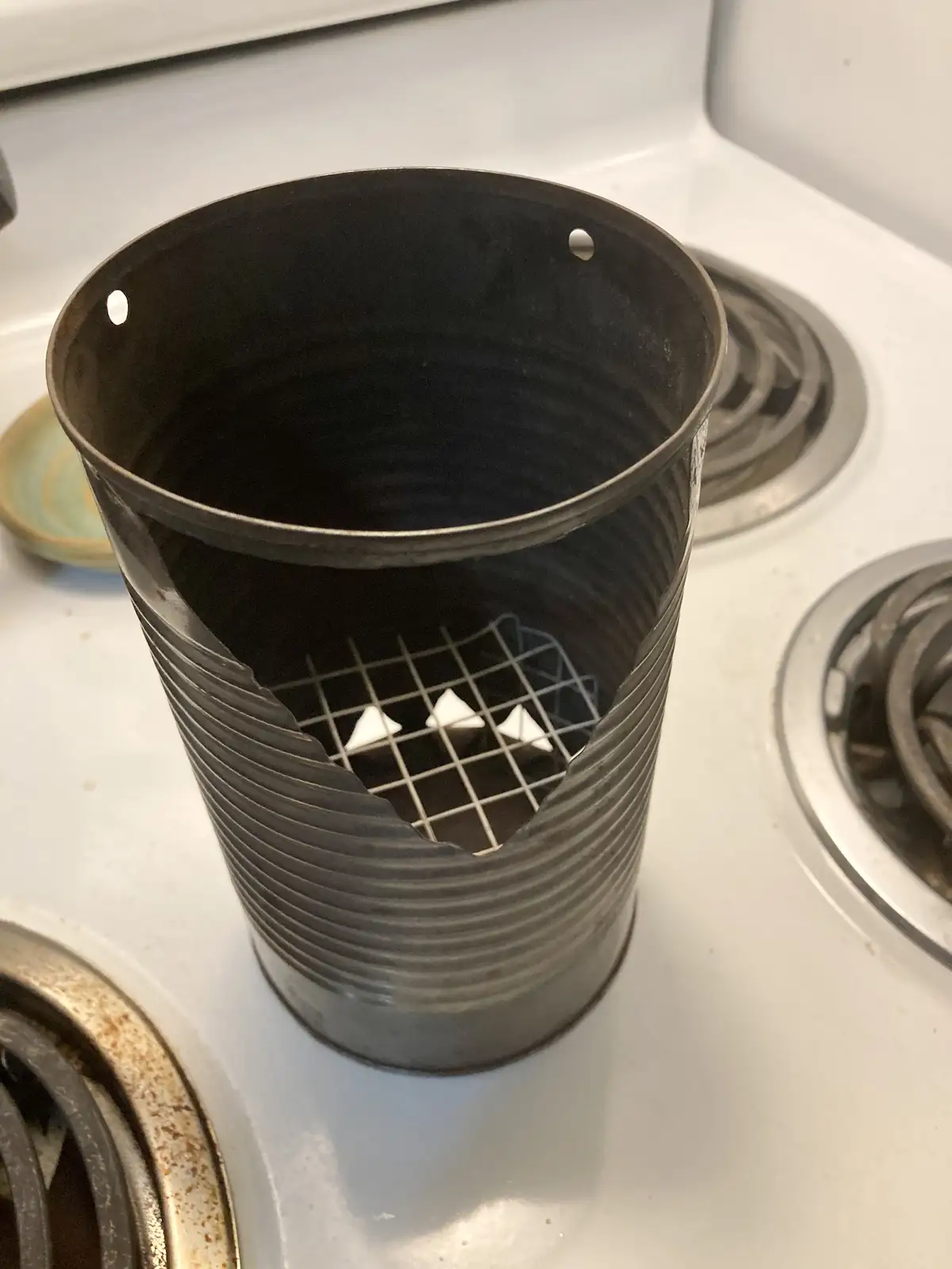 A dark, ribbed metal can sits on a white stovetop, with a wire grid inside holding three white triangular objects. The can…