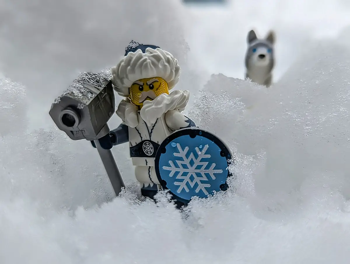 A LEGO minifigure dressed as a winter warrior, with a white beard, holding a frost-covered hammer and a blue shield with a…