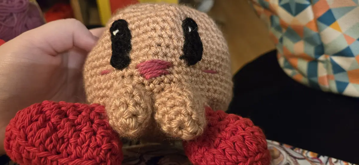 A close-up photo shows a hand holding a beige crocheted plush toy with large black eyes and a pink stitched mouth, its red…