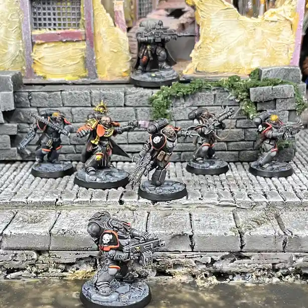 A photo of six painted miniature sci-fi soldiers in grey, orange, and black armor standing on a stone-paved diorama with a…