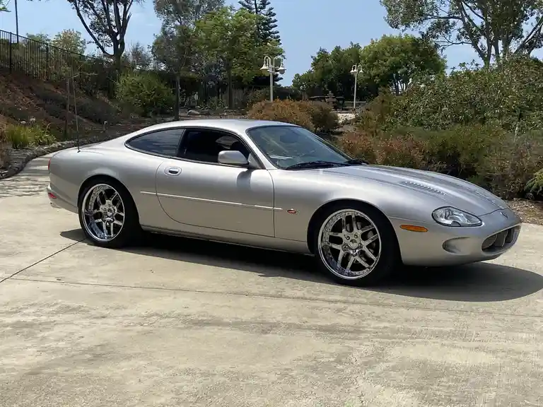 A silver Jaguar XJ220 sports car is parked on a concrete driveway surrounded by greenery and trees under a clear blue sky…