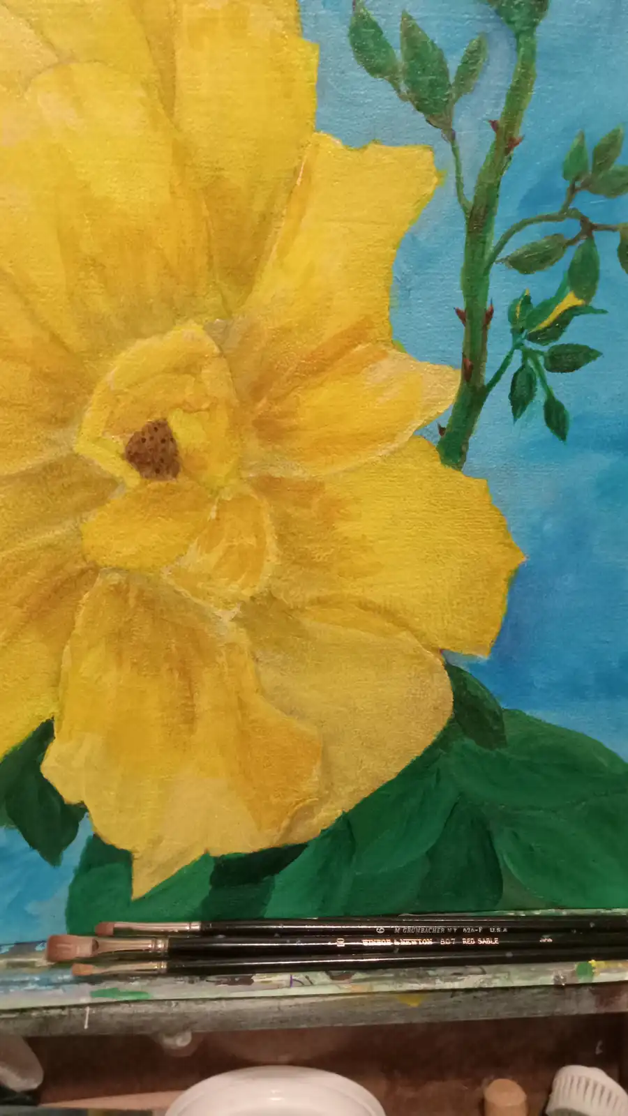 A close-up of a painting shows a large yellow flower with green leaves and stem against a blue background, with paintbrushes…