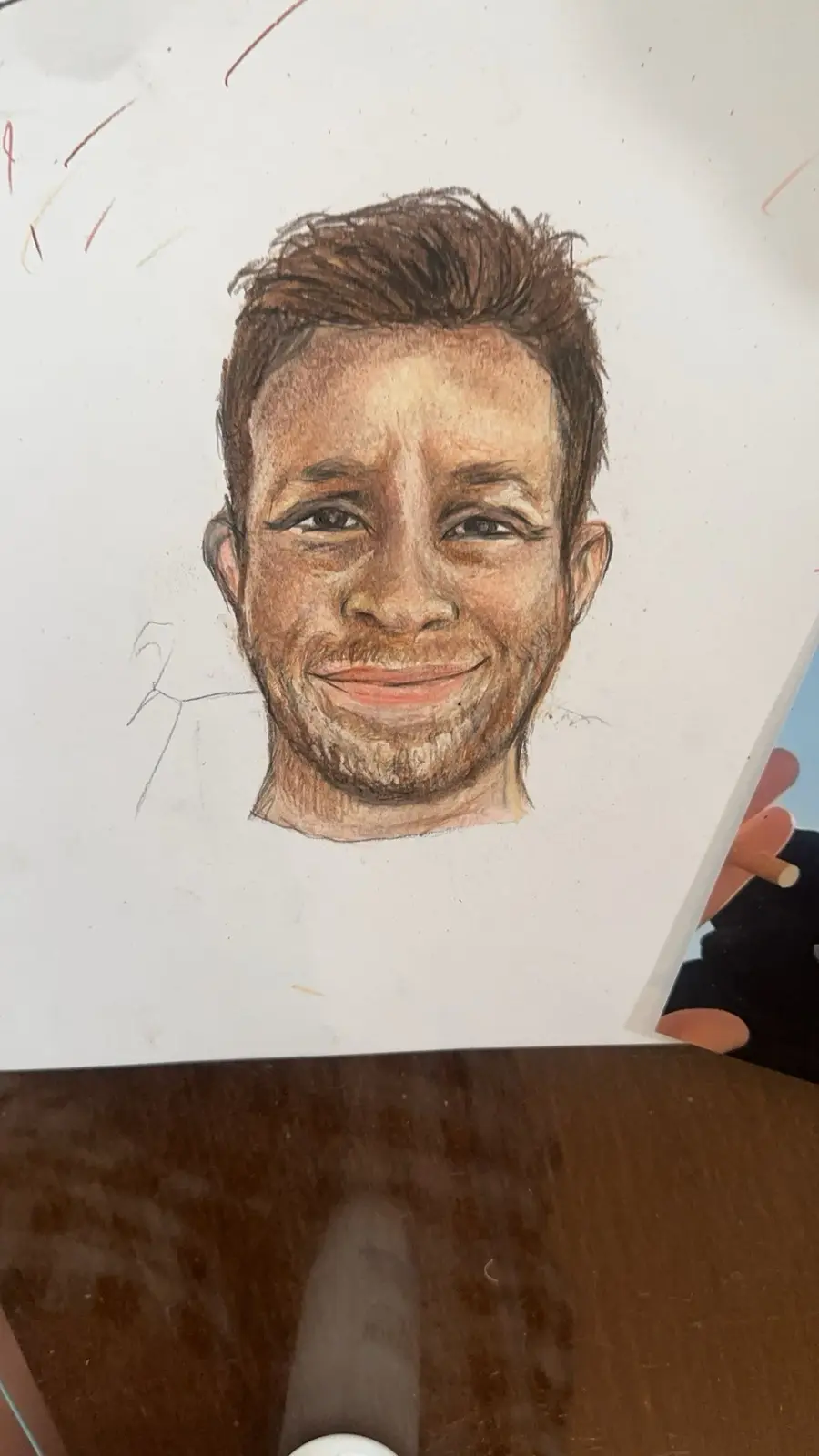 A hand-drawn colored pencil portrait of a smiling man with short brown hair and stubble, shown on a white sheet of paper…