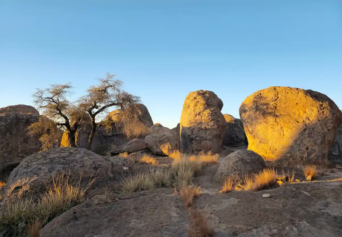 A scenic photo of large, rounded boulders and dry grasses bathed in golden sunlight against a clear blue sky, with a…