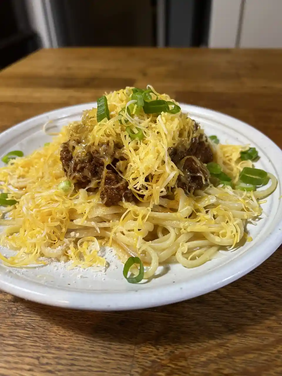 A close-up photo shows a white plate of pasta topped with a meat sauce, a heavy layer of shredded yellow cheese, and chopped…