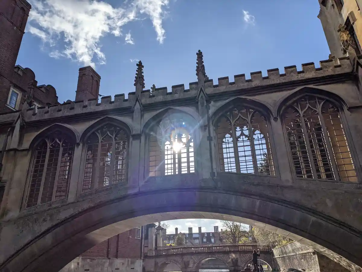A low-angle photo captures the historic Bridge of Sighs in Cambridge, England, with sunlight streaming through its Gothic…