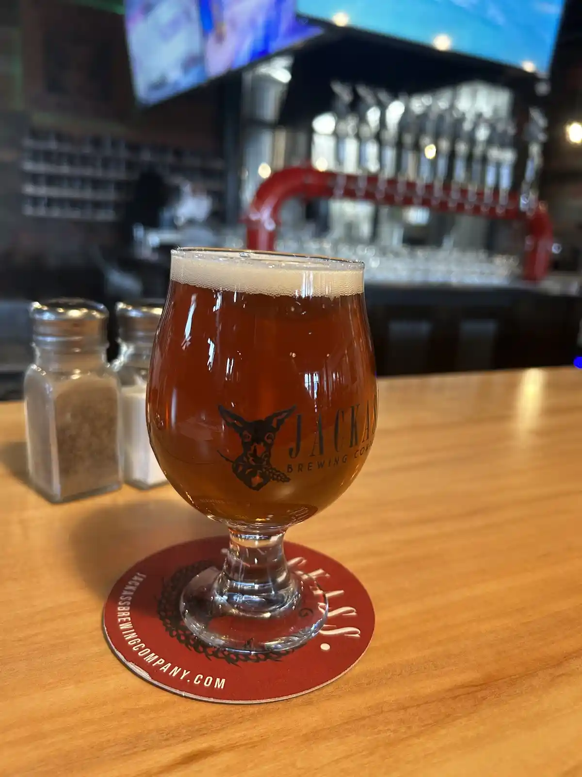 A glass of amber beer with a frothy head sits on a red coaster branded with 'jackassbrewingcompany.com' on a wooden table in…