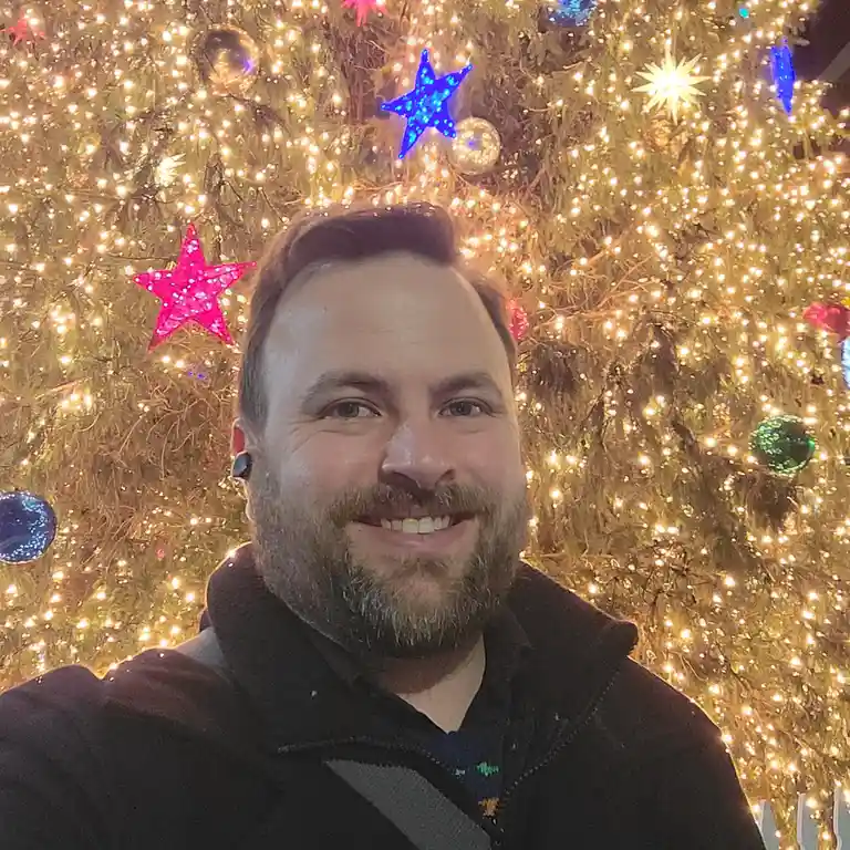 A smiling man with a beard and earbud stands in front of a brightly lit Christmas tree adorned with twinkling lights, pink…
