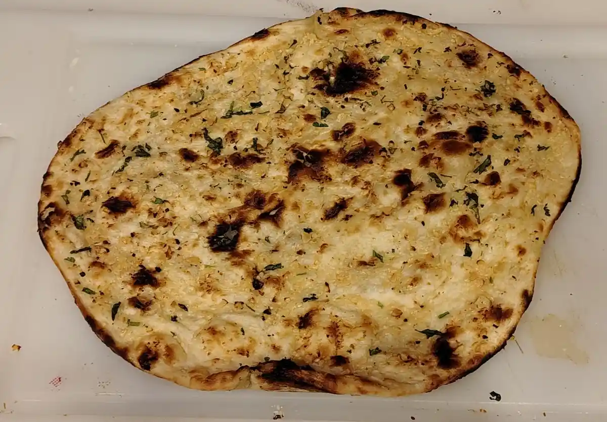 a close-up photo of a round, slightly charred naan bread with green herbs sprinkled on top, resting on a white plastic…