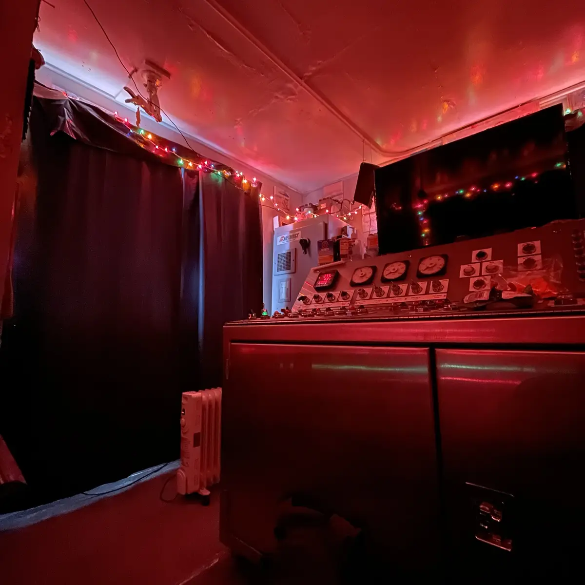 A dimly lit room bathed in red light, featuring a large control panel with dials and buttons, string lights along the…