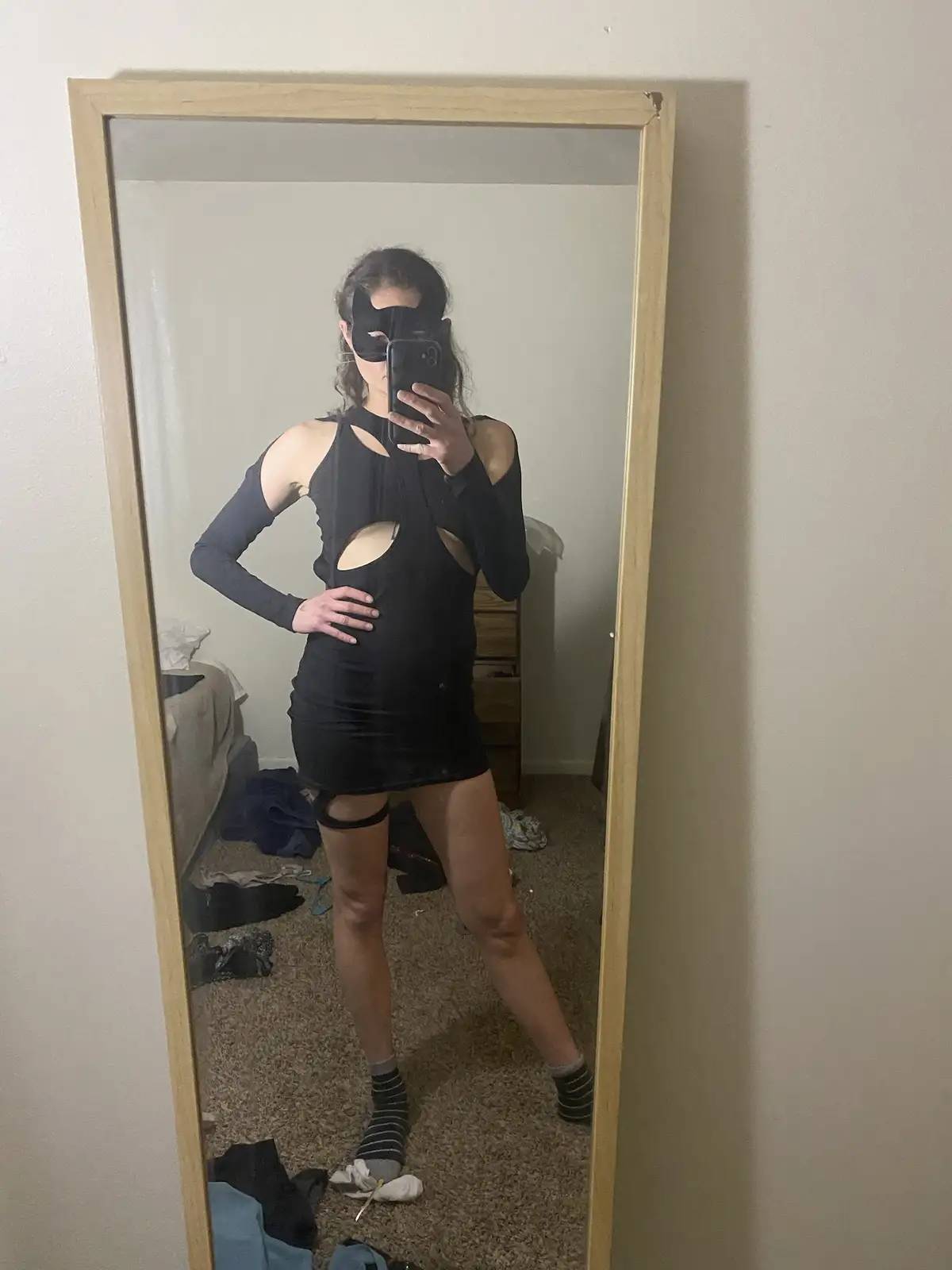 A person wearing a black cat mask and a revealing black dress with cutouts and long sleeves takes a mirror selfie in a…