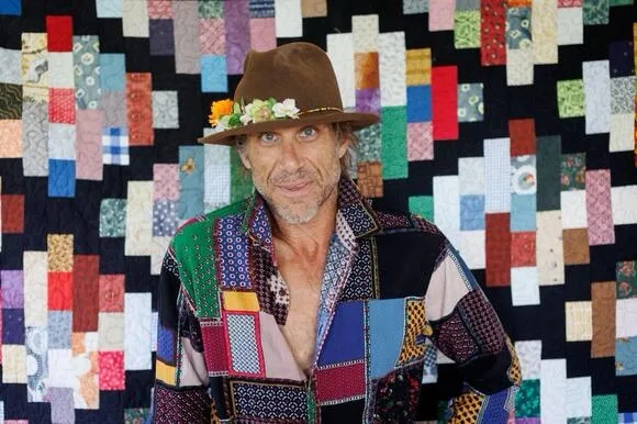 a man with a brown hat adorned with small flowers stands in front of a colorful patchwork quilt background. He wears a…