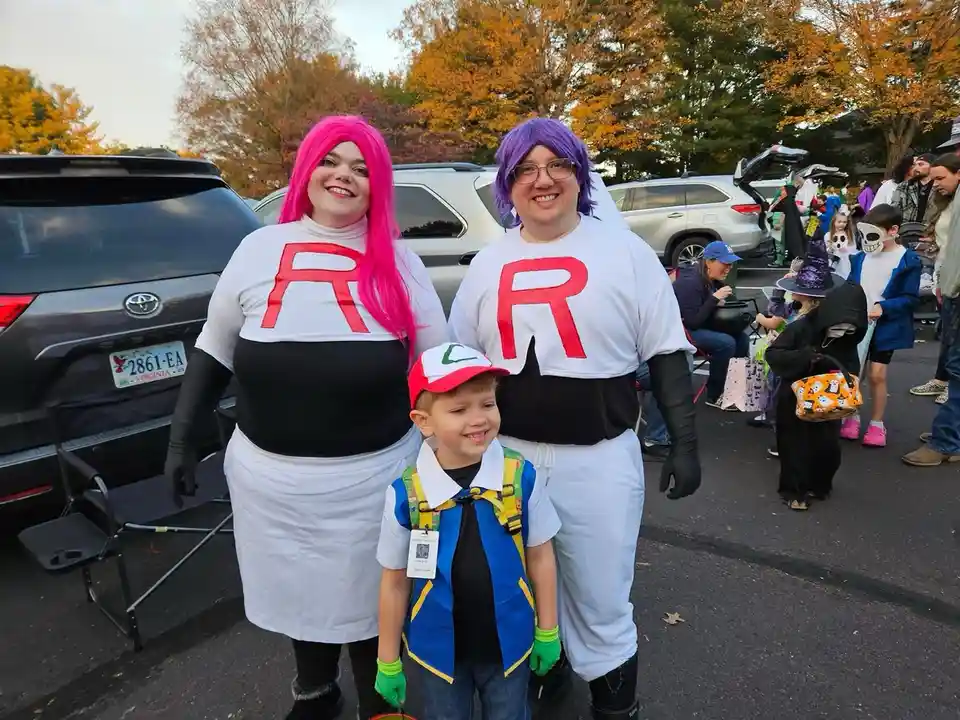 Two adults dressed as Team Rocket villains from Pokémon, wearing white and black outfits with large red 'R' logos and…