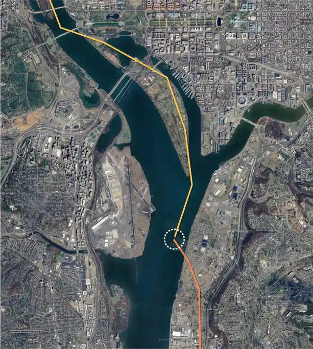 An aerial satellite image of a city with a river, showing two orange lines: one long yellow line crossing the river and a…