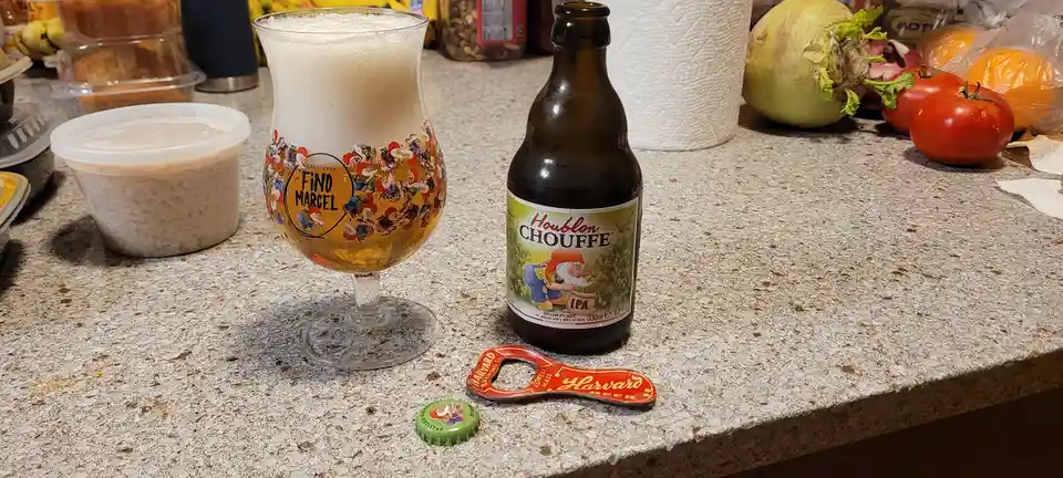 A glass of Chouffe Houblon beer with a thick white head sits next to its bottle on a kitchen counter, the glass features a…