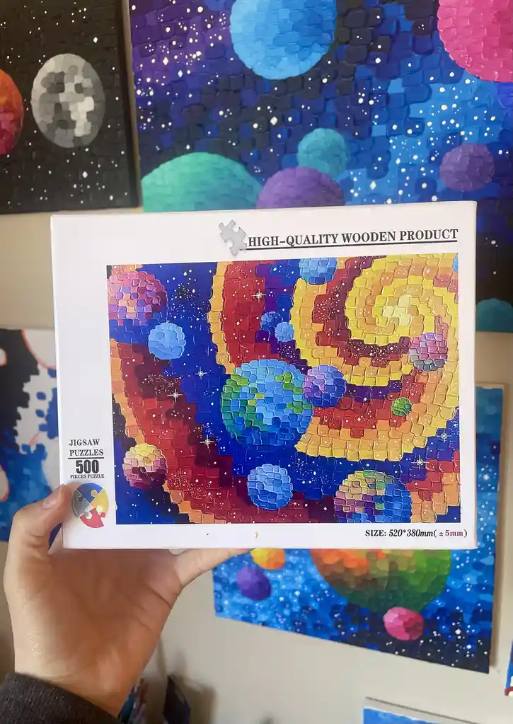 A hand holds a 500-piece jigsaw puzzle box featuring a colorful, pixelated space scene with planets and stars. The box text…