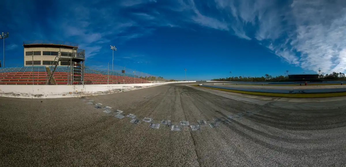 A wide-angle photo of an empty race track with checkered flag markings at the finish line, red and blue bleachers, and a…