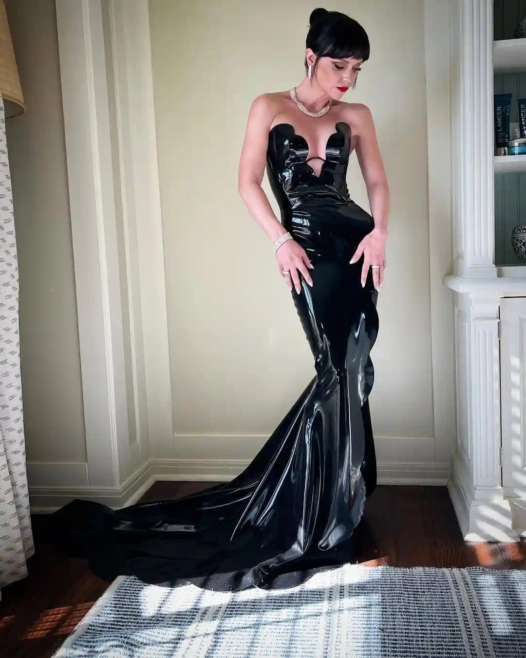 A woman with dark hair and red lipstick stands in a room wearing a long, shiny black latex gown with a plunging neckline and…