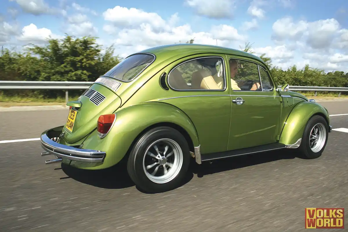 A lime-green classic Volkswagen Beetle is shown driving on a road under a partly cloudy sky, with a man visible in the…