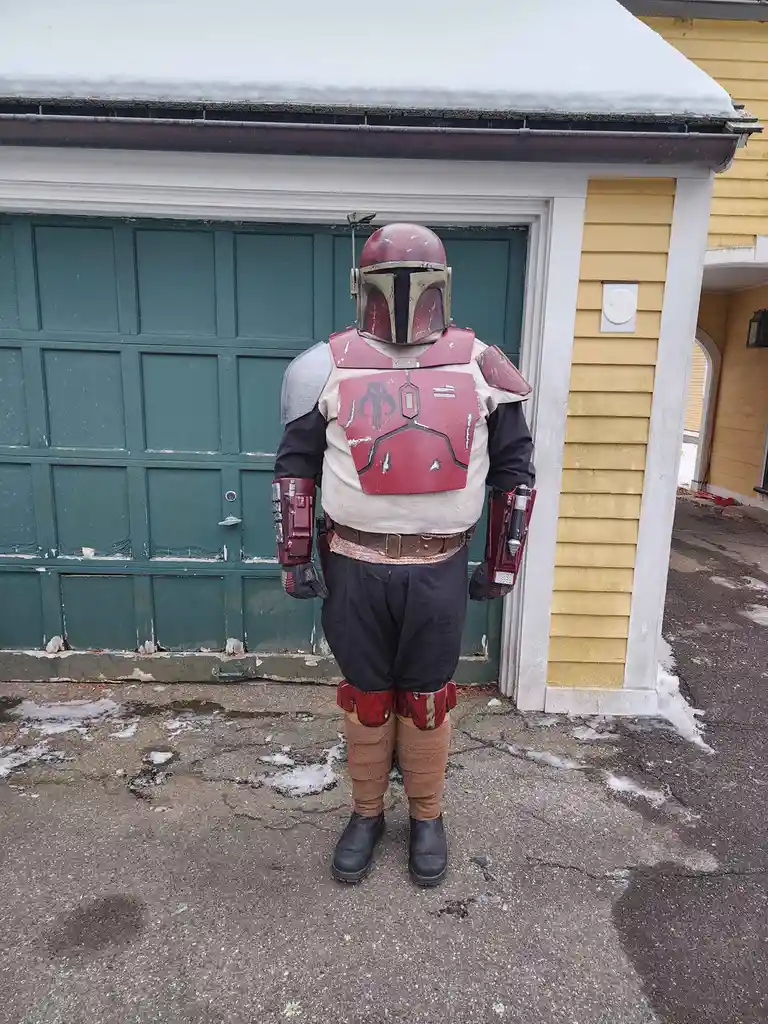 A person in a detailed Boba Fett costume stands with their back to the camera in front of a green garage door and yellow…