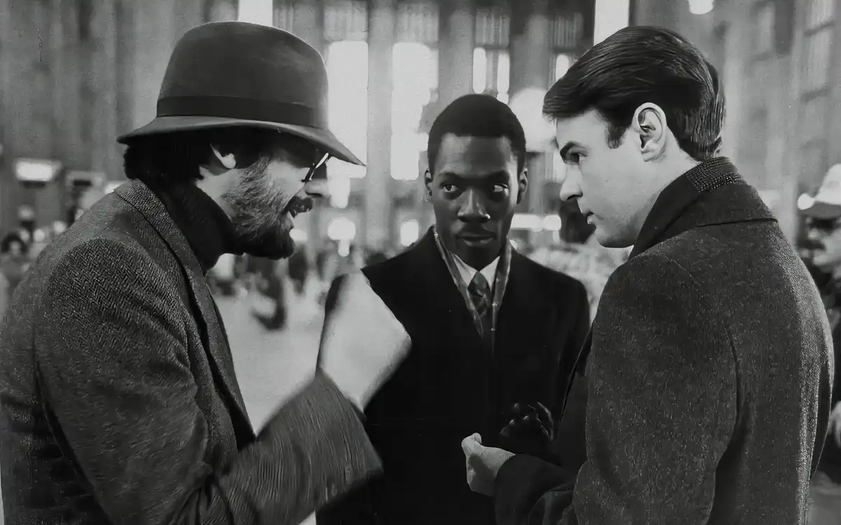 A black-and-white photo shows three men in suits in what appears to be a public space. The man on the left, wearing a fedora…