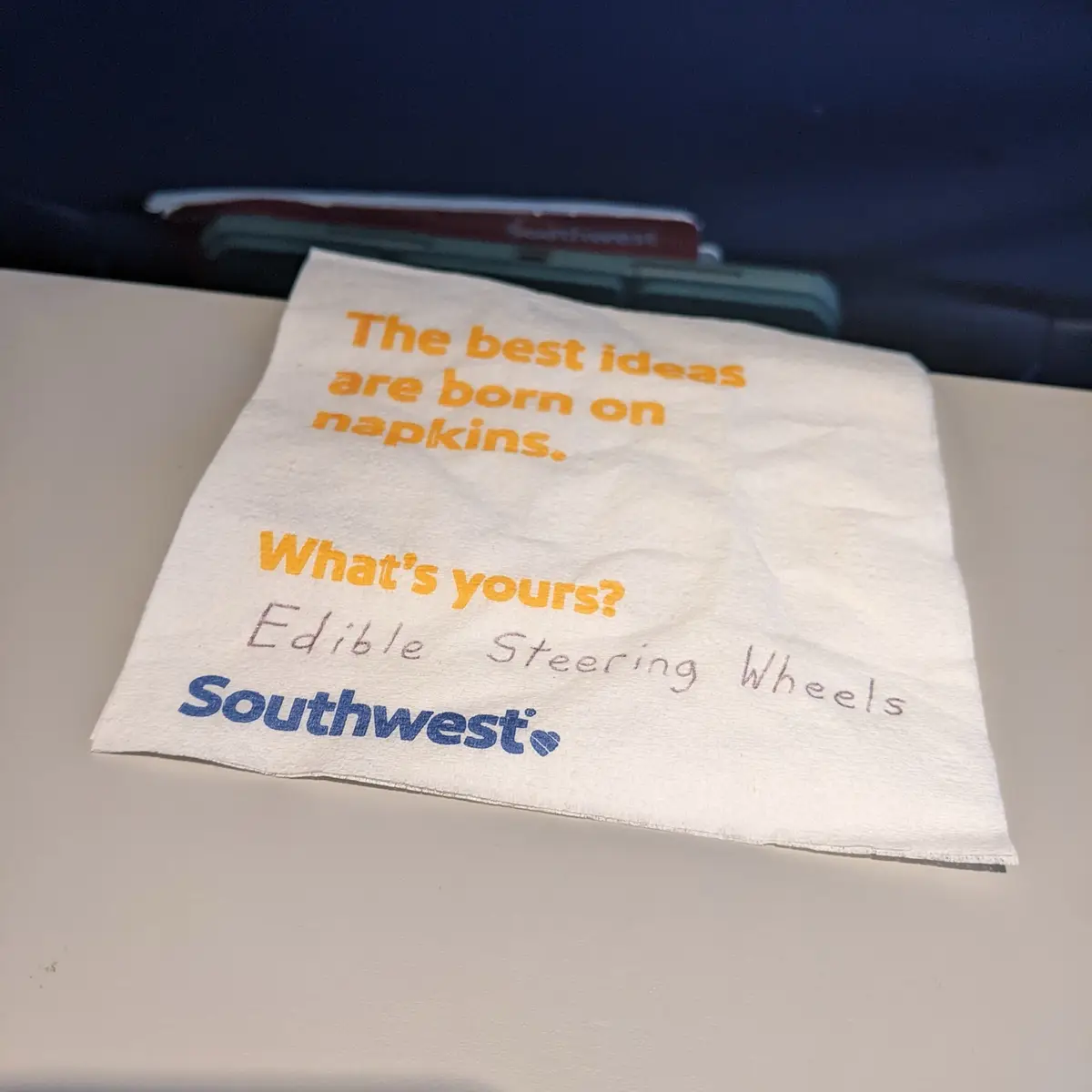 A close-up photo of a Southwest Airlines branded napkin on an airplane tray table. The napkin has printed orange text…