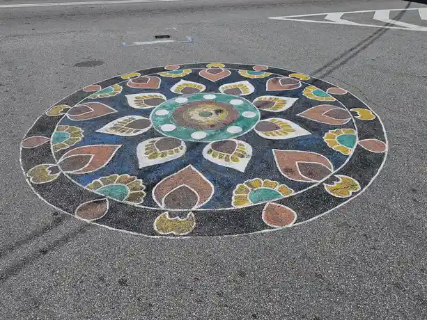 A colorful, intricate mandala painted on asphalt pavement, featuring concentric circles and symmetrical floral patterns in…