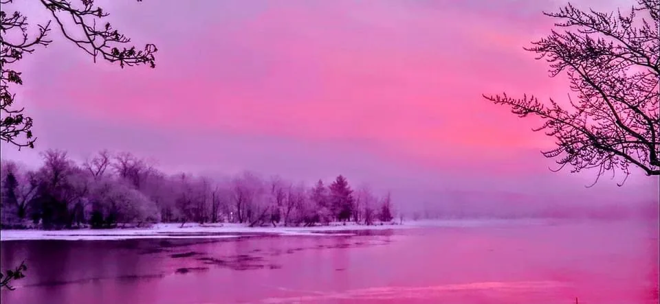 a serene winter landscape with a frozen lake, snow-covered trees, and bare branches framing the scene, all bathed in an…