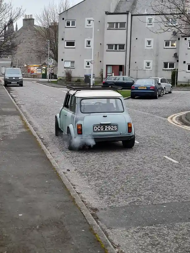 A light blue classic Mini car with a roof rack is driving down a residential street, emitting thick smoke from its exhaust…