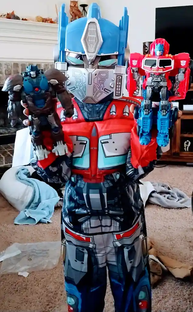 A child wearing a full Optimus Prime costume stands indoors holding two different Optimus Prime action figures, one in each…