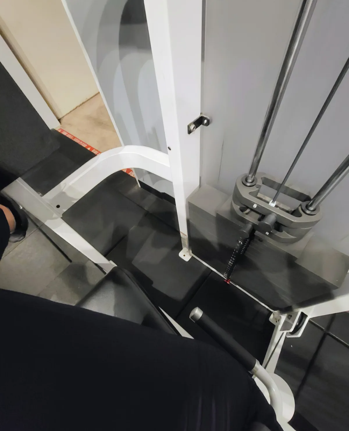 A close-up photo of a person sitting on a gym machine, showing the machine's black padded seat and white frame, with a red…