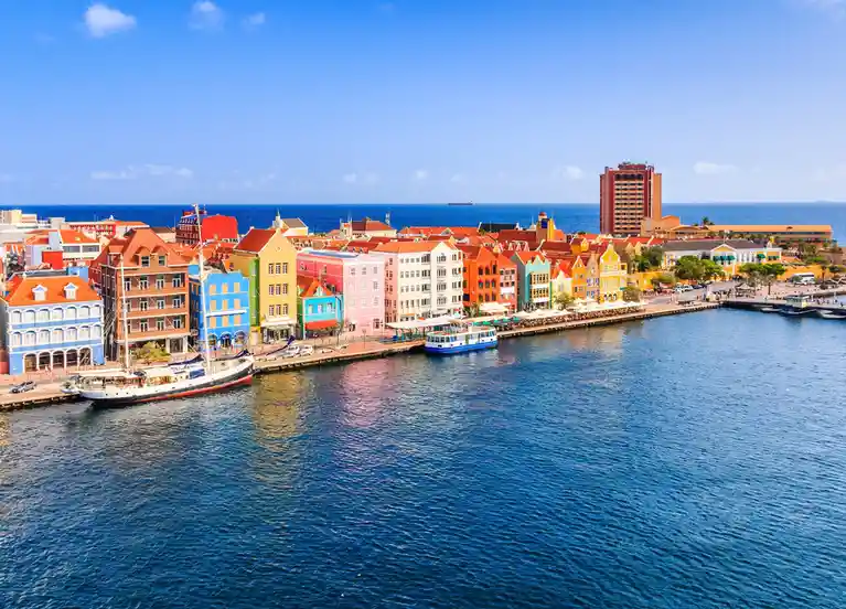 A vibrant, high-angle photograph of a waterfront city with brightly colored buildings lining a harbor under a clear blue…