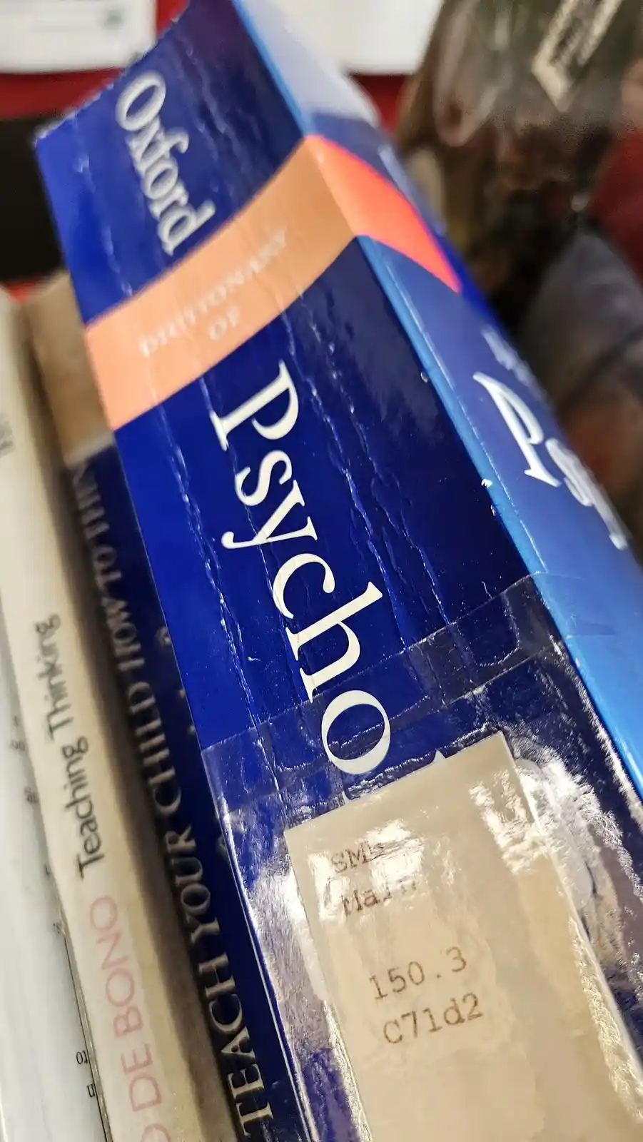 A close-up shot of a blue 'Oxford Dictionary of Psychology' book spine, showing a library sticker with 'SMS', '150.3', and…