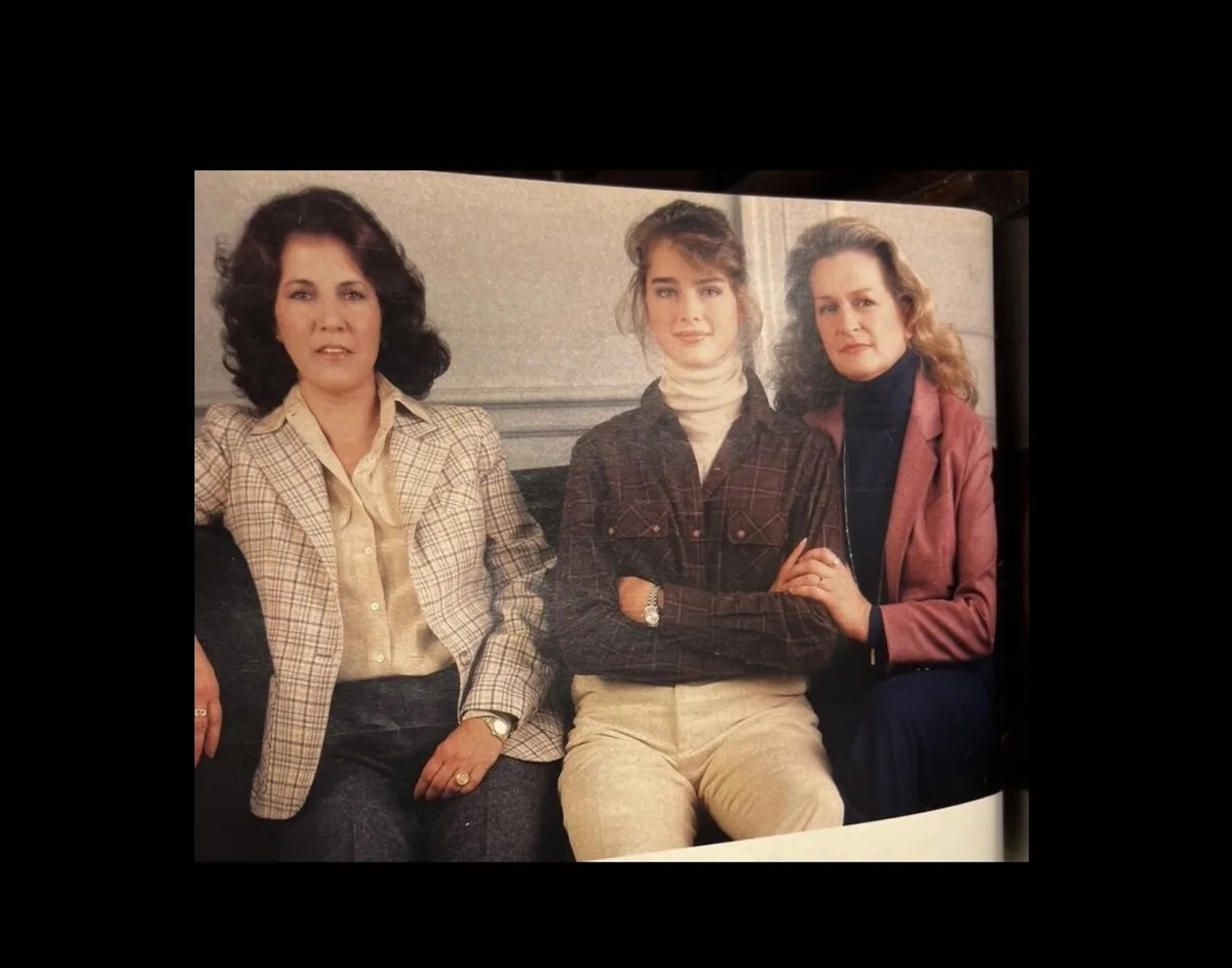 A vintage photo shows three women sitting side-by-side on a couch, dressed in 1980s-style clothing including plaid blazers…